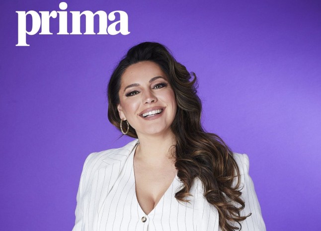 Kelly Brook is wearing a white suit and she's smiling. She is standing in front of a purple background.