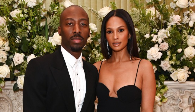 Alesha Dixon and Akuza Ononye at a red carpet event. Akuza is wearing a black suit while Alesha is wearing a black dress. They are posing with their arms around each other in front of a white floral backdrop