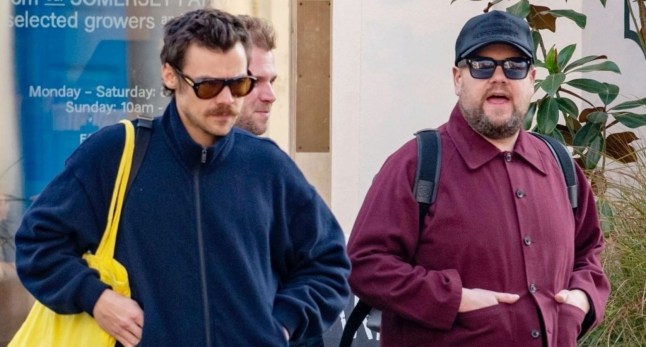 Harry Styles wears a blue tracksuit and yellow tote bag as he walks next to James Corden (R), both have hands in their pockets.