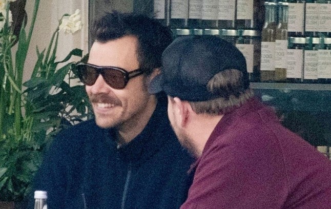 Harry Styles (L) grins as he sits next to James Corden outside cafe