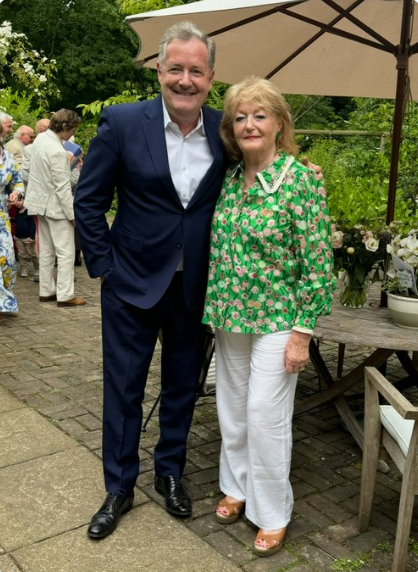 Piers Morgan in a navy suit poses with his mother Gabriella in a green floral blouse and white trousers outside