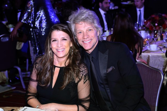 Dorothea Hurley and Jon Bon Jovi posing at dinner table during event. He wears black suit and tie and she wears black dress.
