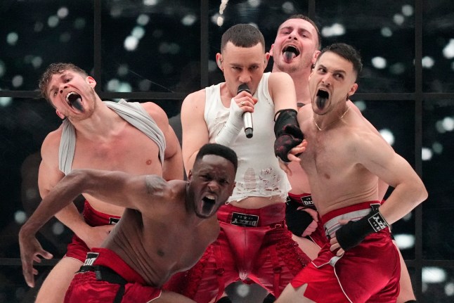Olly Alexander in a white top and red trousers holding a microphone with four topless back up dancers surrounding him