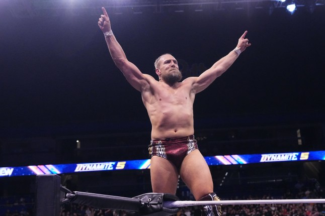 A topless Bryan Danielson holding his arms up while in the ringagainst Lee South Kazuchika Okada
