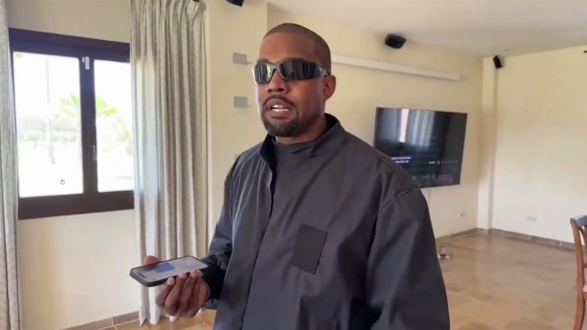 Kanye West holds phone as he speaks on Twitch in empty room, with TV behind him. He wears sunglasses and black jacket.