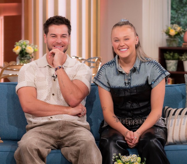 JoJo Siwa and Chris Hughes sitting on a sofa together in casual clothing smiling at the camera