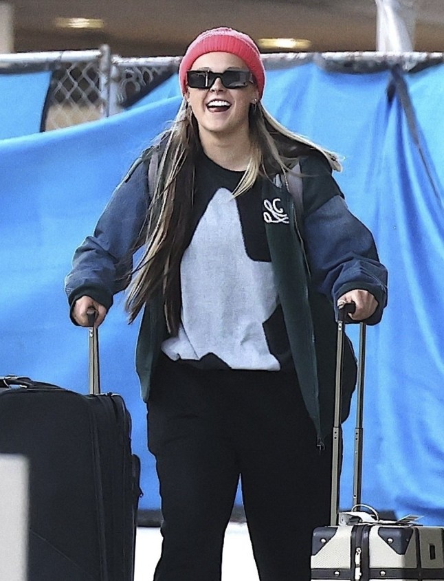 JoJo Siwa wearing a grey and black top, jacket, pink beanie, sunglasses, black trousers rolling a pair of suitcases as she smiles