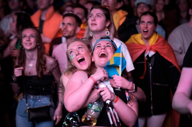 Two Eurovision fans with Swedish flags smiling and hugging with their faces lit up by a screen at a Eurovision watch party.