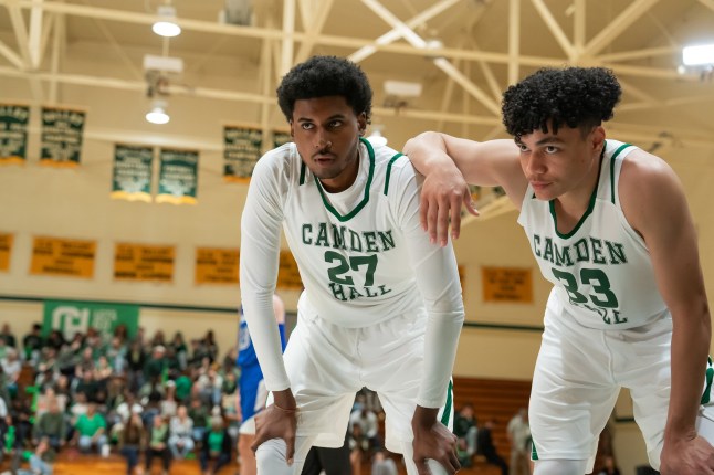 Two teen boys in white and green basketball uniform lean on each other while bent over on the court